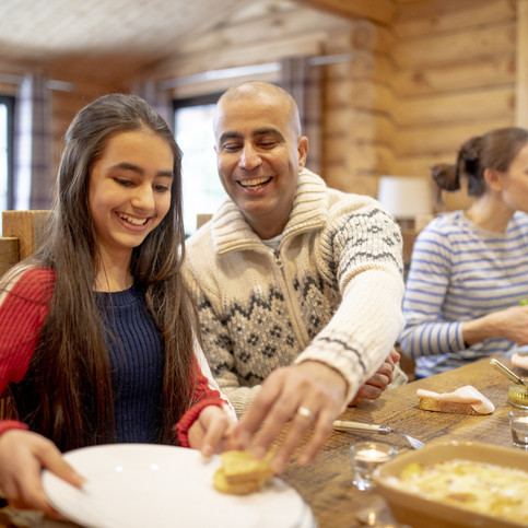 A mid adult man serves his daughter dinner in a log cabin whilst his wife prepares to serve lettuce.